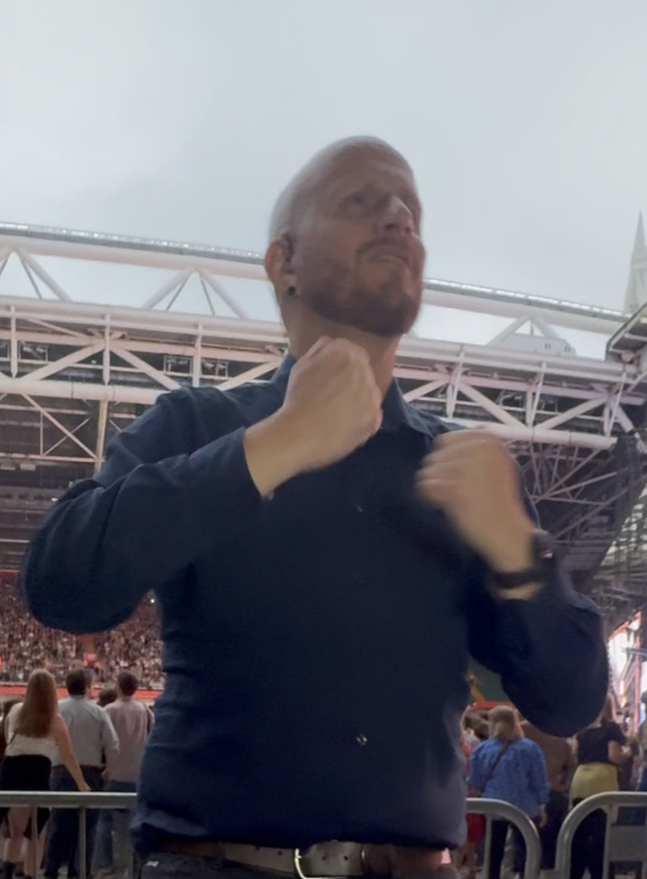 Adrian Bailey signing in British Sign Language at an outdoor stadium concert, with the crowd and stage barriers behind him